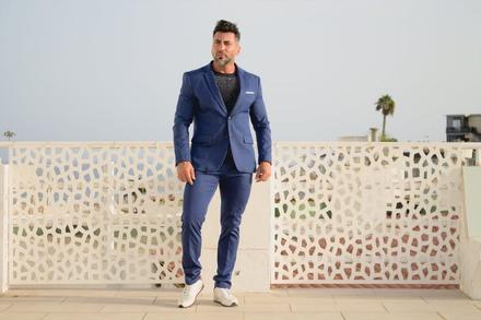 Pablo de cuerpo entero con traje azul, de pie ante una celosía blanca frente al mar.