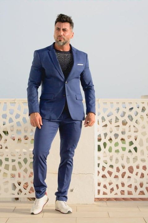 Pablo de cuerpo entero con traje azul, de pie ante una celosía blanca frente al mar.