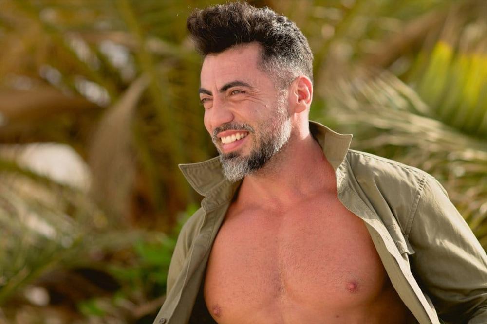 Pablo sonriendo con una camisa caqui abierta, entre palmeras.