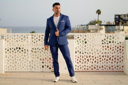 Pablo de cuerpo entero con traje azul, ajustándose la chaqueta, con el mar al fondo.